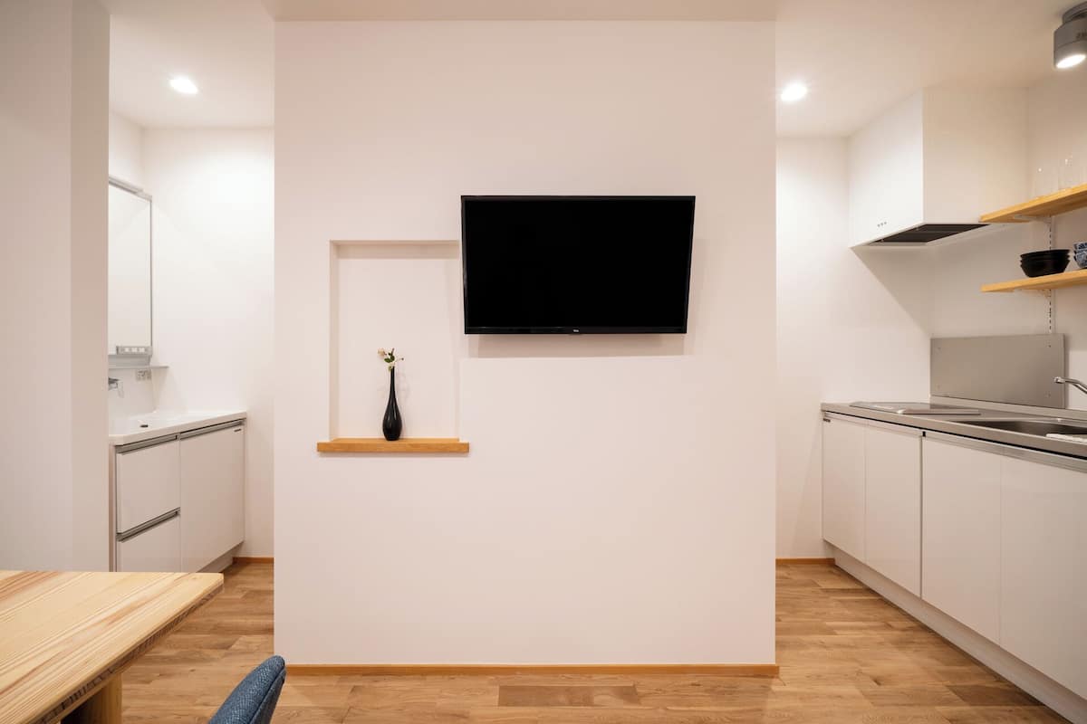 Une cuisine moderne avec des meubles blancs, un plan de travail en bois, un vase noir posé sur une étagère et un téléviseur à écran plat fixé au mur.