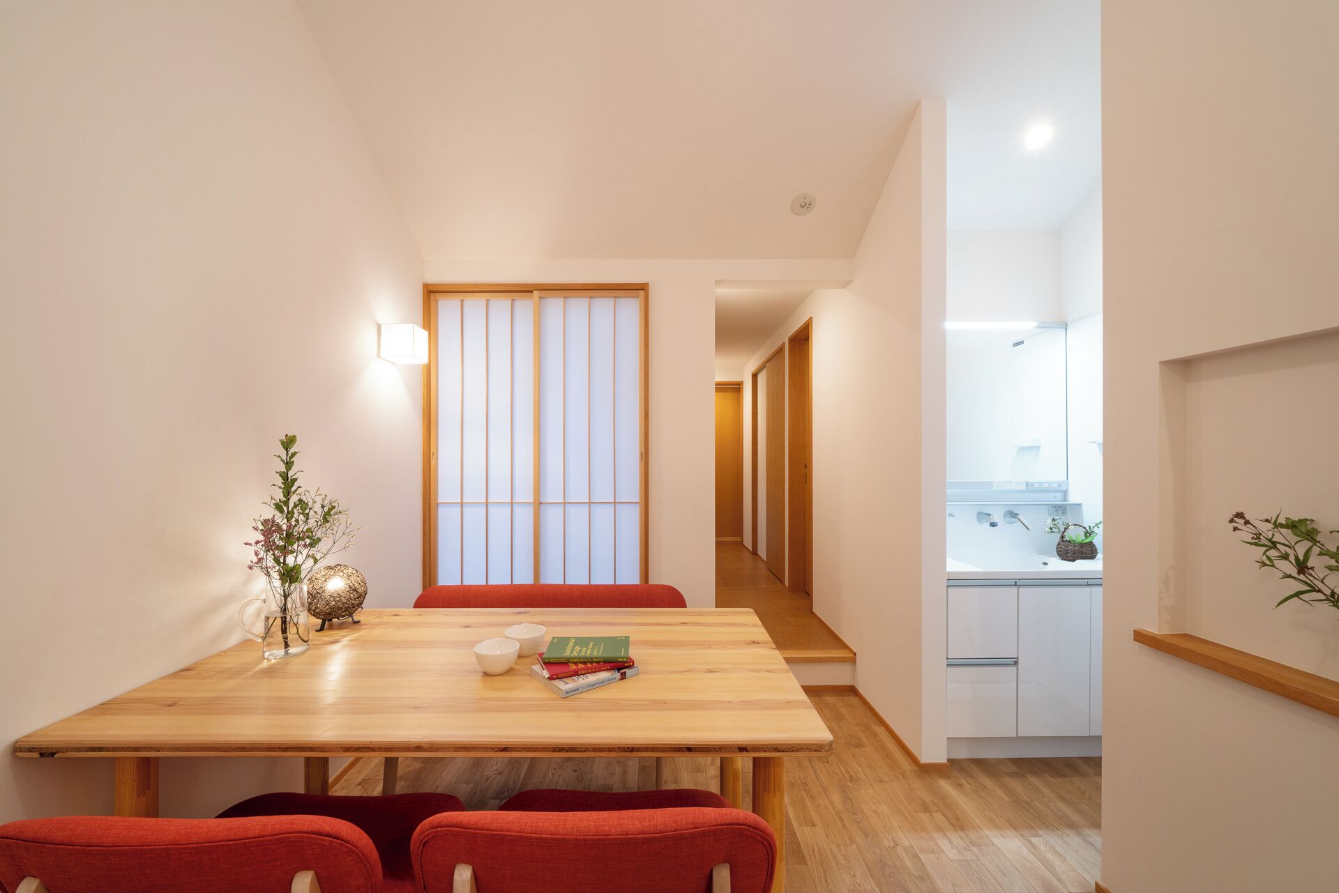 A modern interior with a wooden dining table, red chairs, a sliding shoji screen, and a view into a kitchen area.