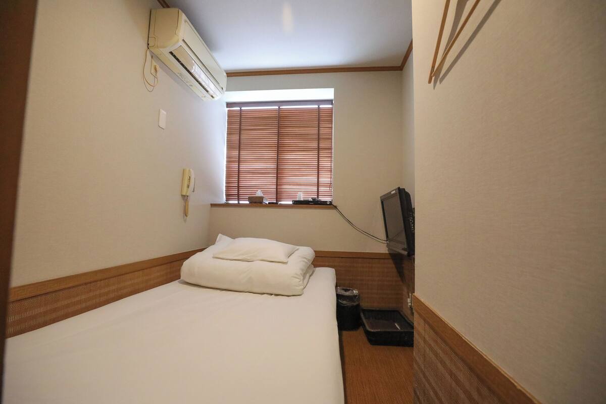 A hotel room with a bed, a window with blinds, an air conditioning unit, a television, and a small trash bin.