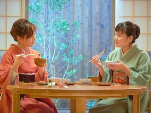 To personer sidder ved et rundt træbord med traditionelle japanske retter, spisepinde og en lille plante i baggrunden.