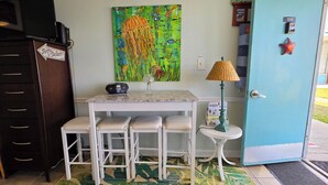 A small kitchenette with a white table, bar stools, a painting, a lamp, and a door with a starfish decoration. - Cozy studio with refreshing AC, pool, and 3-minute walk to the beach (Atlantic Beach)