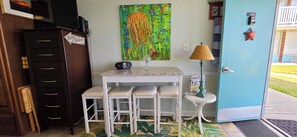 A small kitchenette with a white table, bar stools, a painting, a lamp, and a door with a starfish decoration. - Cozy studio with refreshing AC, pool, and 3-minute walk to the beach (Atlantic Beach)