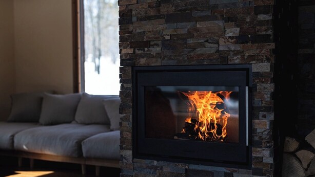 Chimenea de piedra con fuego visible, un sofá y una ventana en una sala de estar.