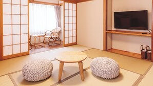 Una habitación japonesa tradicional con suelo de tatami, una mesa de madera baja, dos pufs tejidos, un televisor en un estante y una puerta corredera que da a una ventana con cortina.