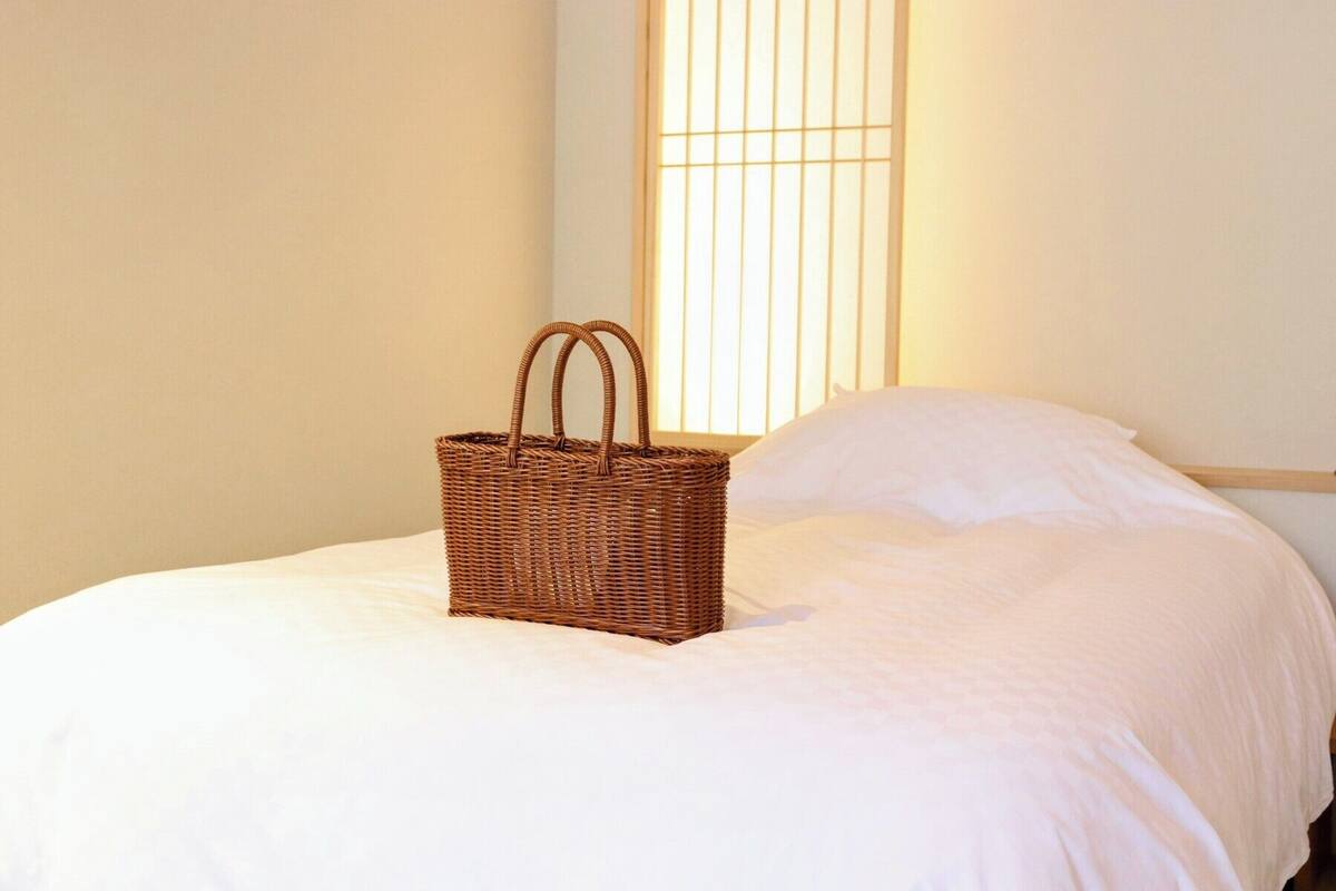 Una cesta de mimbre sobre una cama con ropa blanca y una ventana con persianas verticales al fondo.