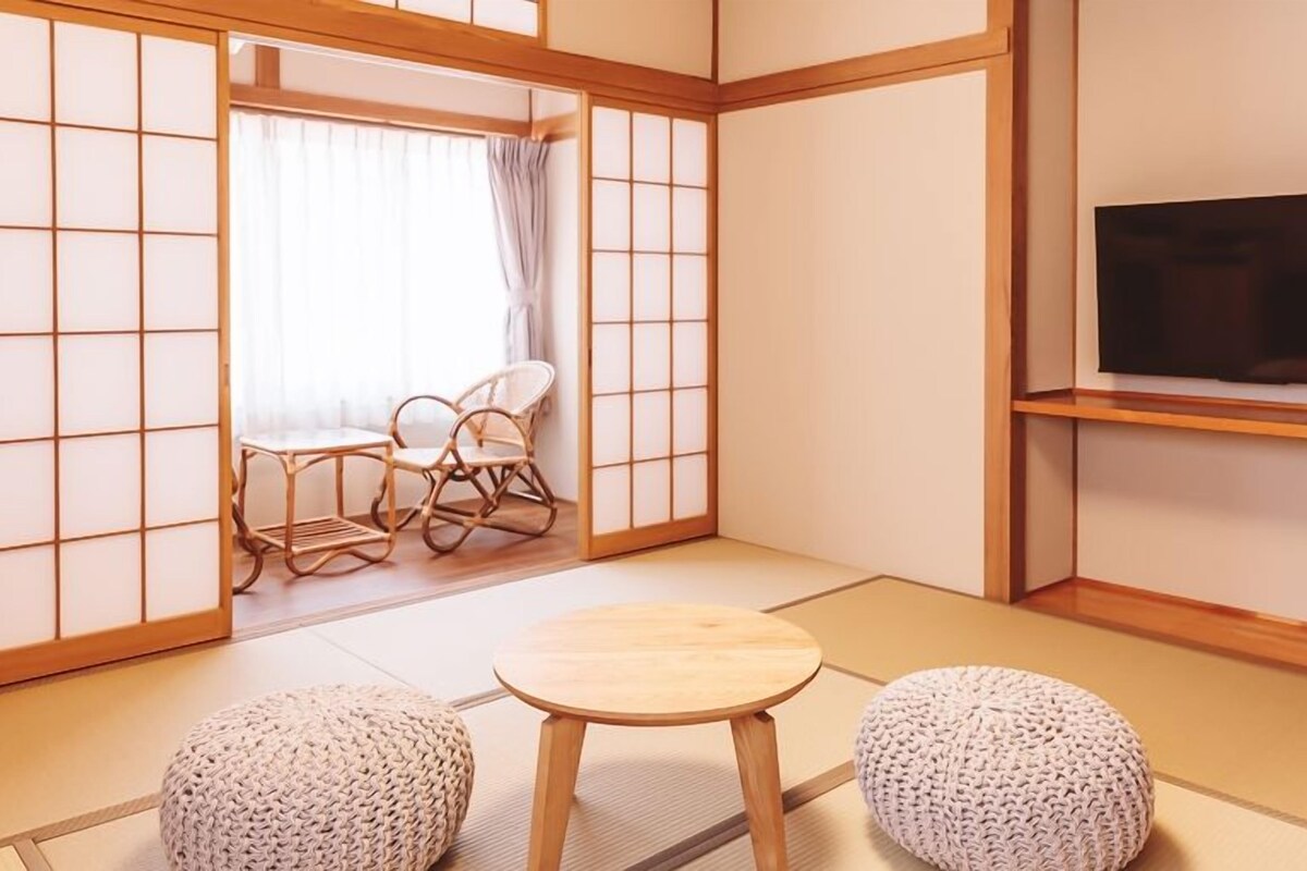 Une pièce de style japonais traditionnel, avec un sol recouvert de tatamis, une table basse, deux sièges rembourrés, un téléviseur posé sur une étagère et une porte coulissante.