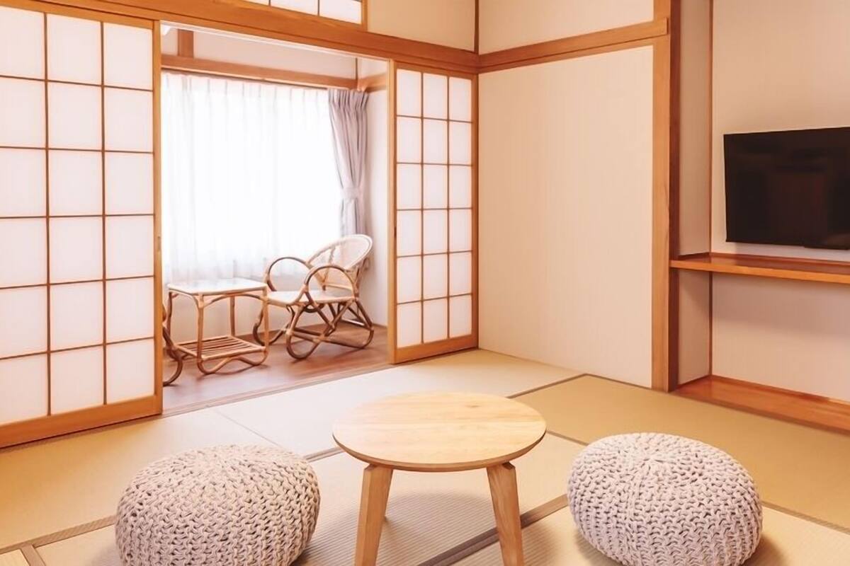 Una habitación de estilo japonés tradicional con suelo de tatami, una mesa baja, dos asientos acolchados, un televisor en un estante y un puertas correderas.