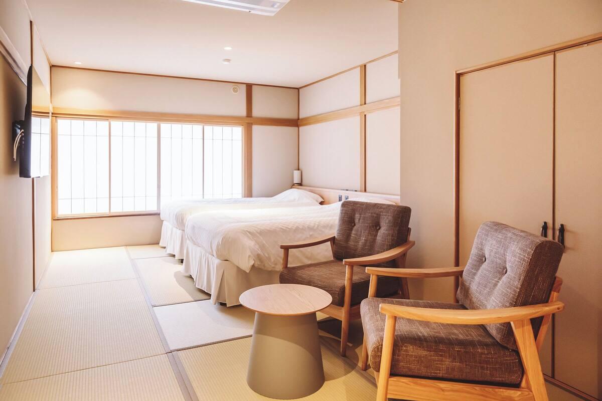 Habitación de hotel con suelo de tatami, una cama, dos sillas, una mesita redonda y un televisor.