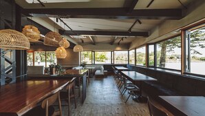 Interior de restaurante con mesas de madera, lámparas colgantes tejidas y amplias ventanas que ofrecen vistas al exterior.