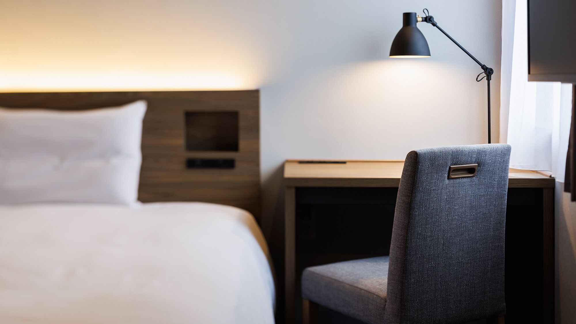 A hotel room with a bed, a desk, a chair, and a desk lamp.