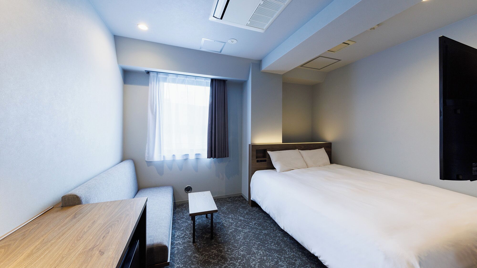 A hotel room with a bed, a desk, a chair, a small table, a window with curtains, and a wall-mounted TV.