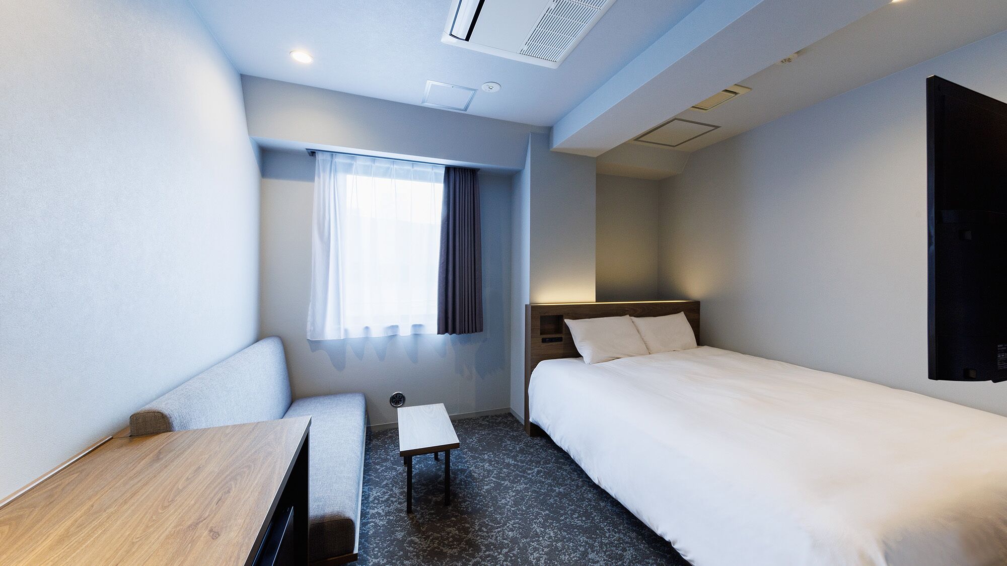A hotel room with a bed, a desk, a chair, a small table, a window with curtains, and a wall-mounted TV.