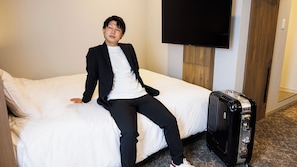Une personne est assise sur un lit dans une chambre d’hôtel, avec une grande valise noire et un téléviseur fixé au mur.