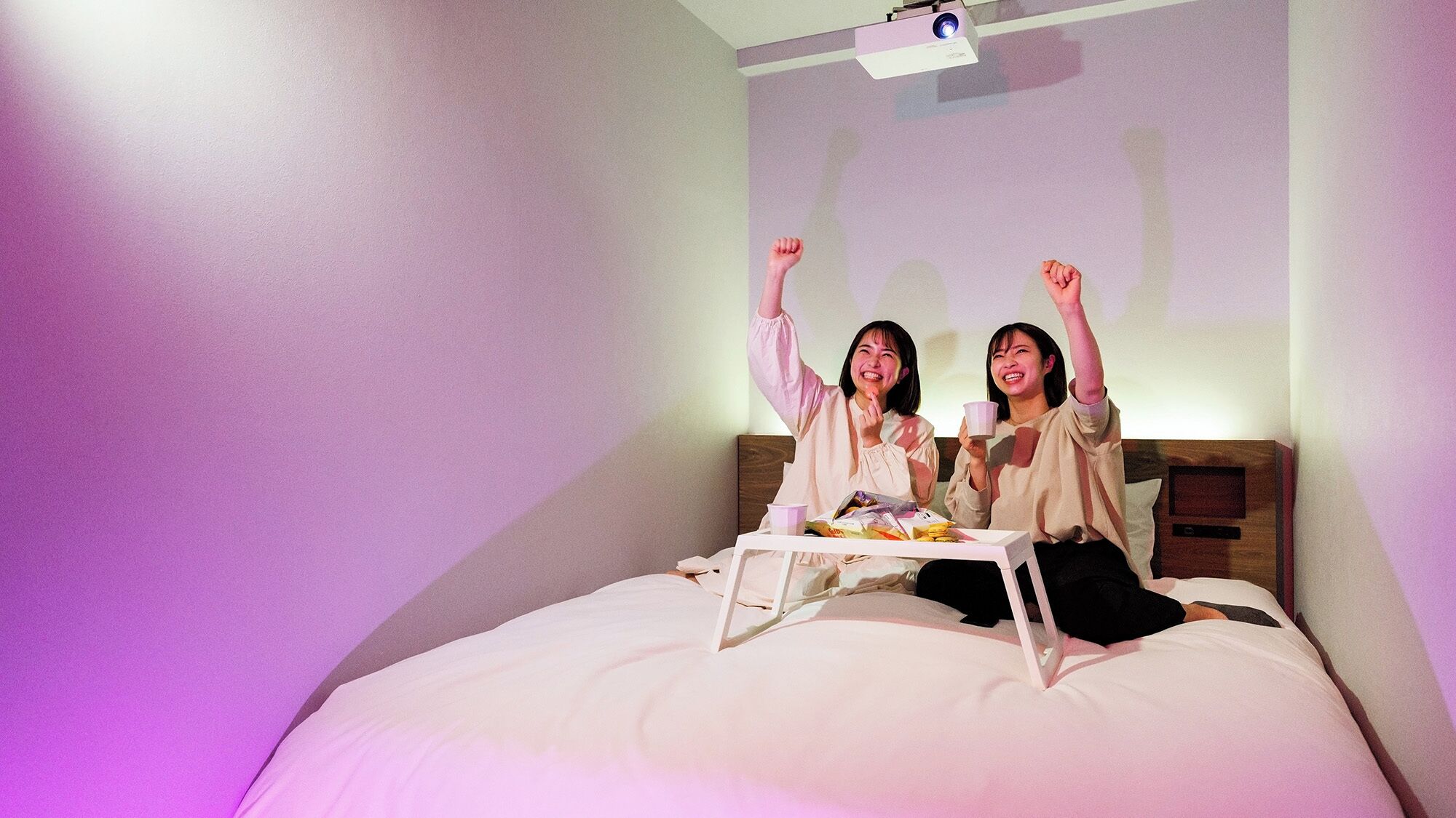 Two people sitting on a bed with a projector screen in the background.