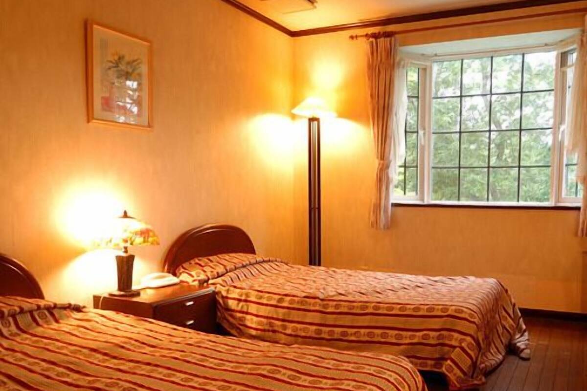 Ein Zimmer mit zwei Betten, einem Nachttisch, einer Lampe, einem Fenster mit Vorhängen und einem gerahmten Bild an der Wand.