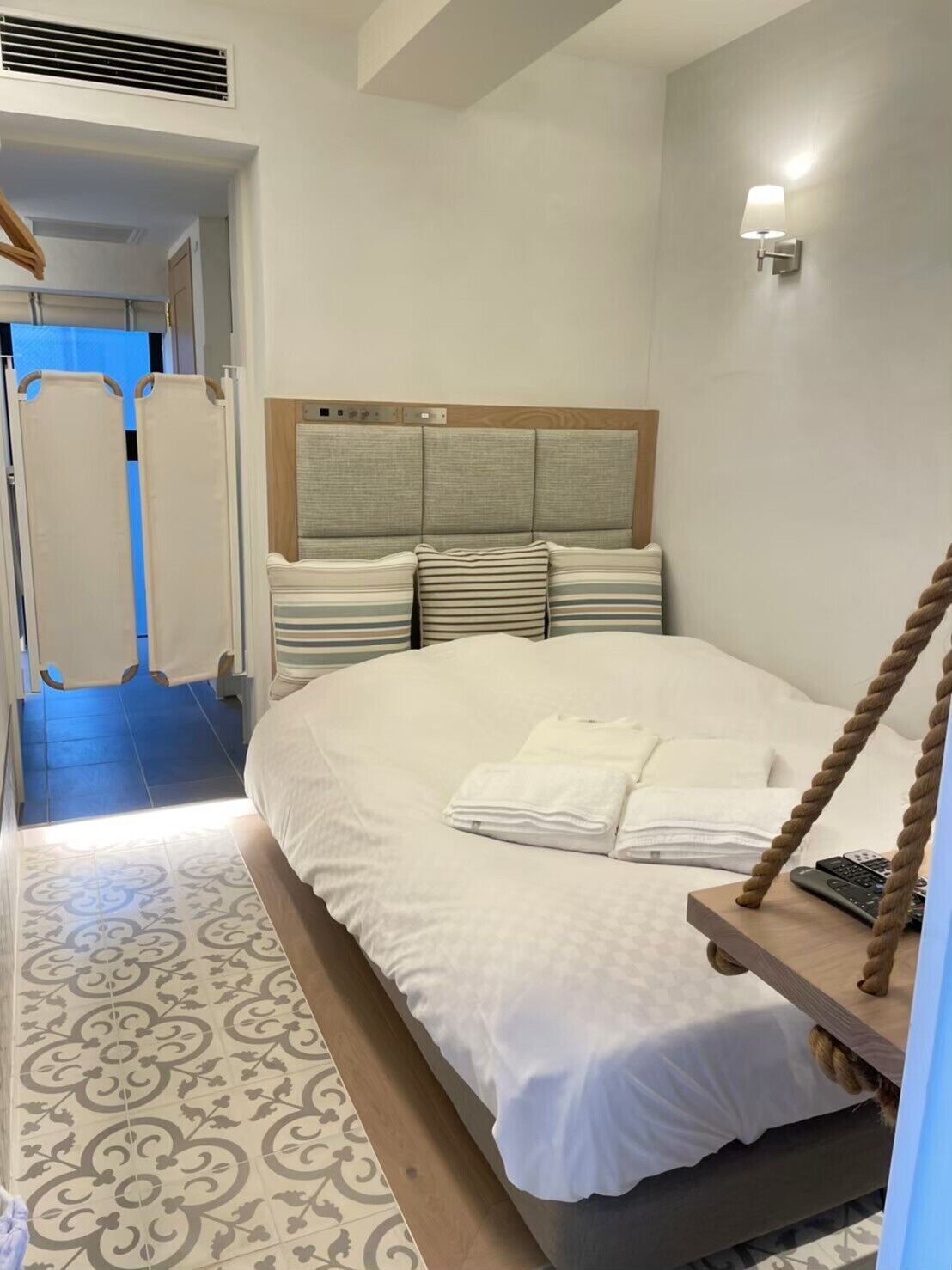 A bedroom with a bed, a headboard, a wall-mounted light, and a wooden bedside table with a remote control.