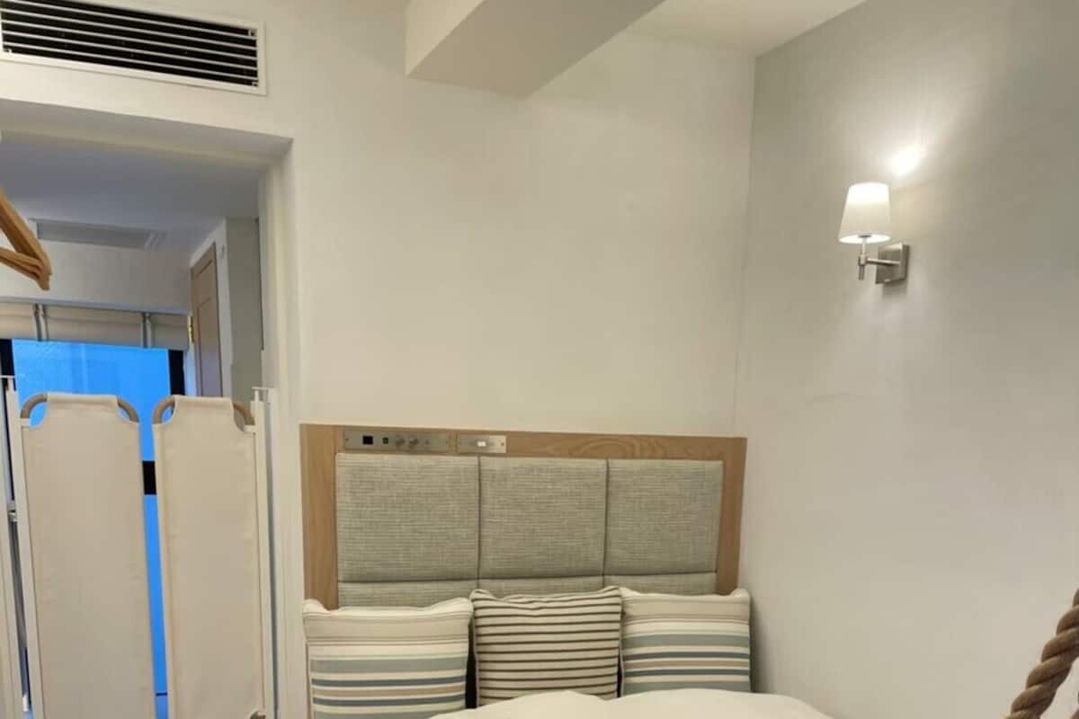 Une chambre à coucher comprenant un lit, une tête de lit, une lampe fixée au mur et une table de chevet en bois avec une télécommande.