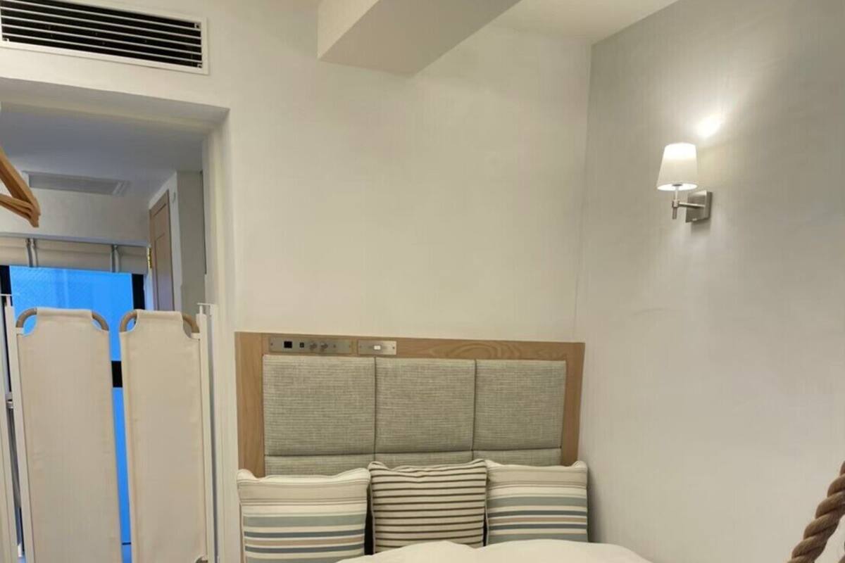 A bedroom with a bed, a headboard, a wall-mounted light, and a wooden bedside table with a remote control.
