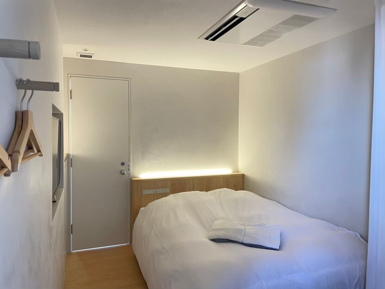 A modern hotel room with a wooden headboard, a bed with white bedding, a door with a light strip, and a wall-mounted towel rack.