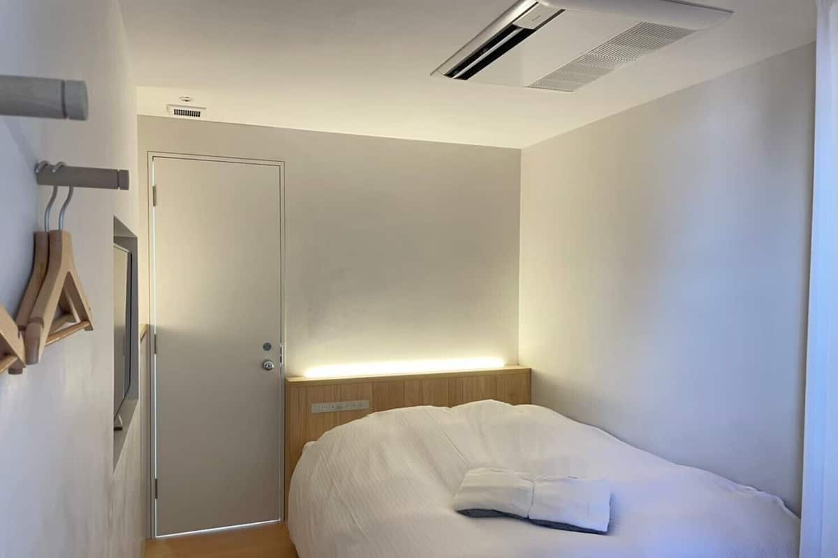 Une chambre d’hôtel moderne avec une tête de lit en bois, un lit avec une literie blanche, une porte dotée d’une bande lumineuse et un porte-serviettes fixé au mur.