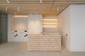 A reception desk with a wooden finish, a glass-top counter, and a modern design. There are white cabinets with geometric cutouts to the left, and a door with a red sign to the right. - Hotel Siro (Tokyo)
