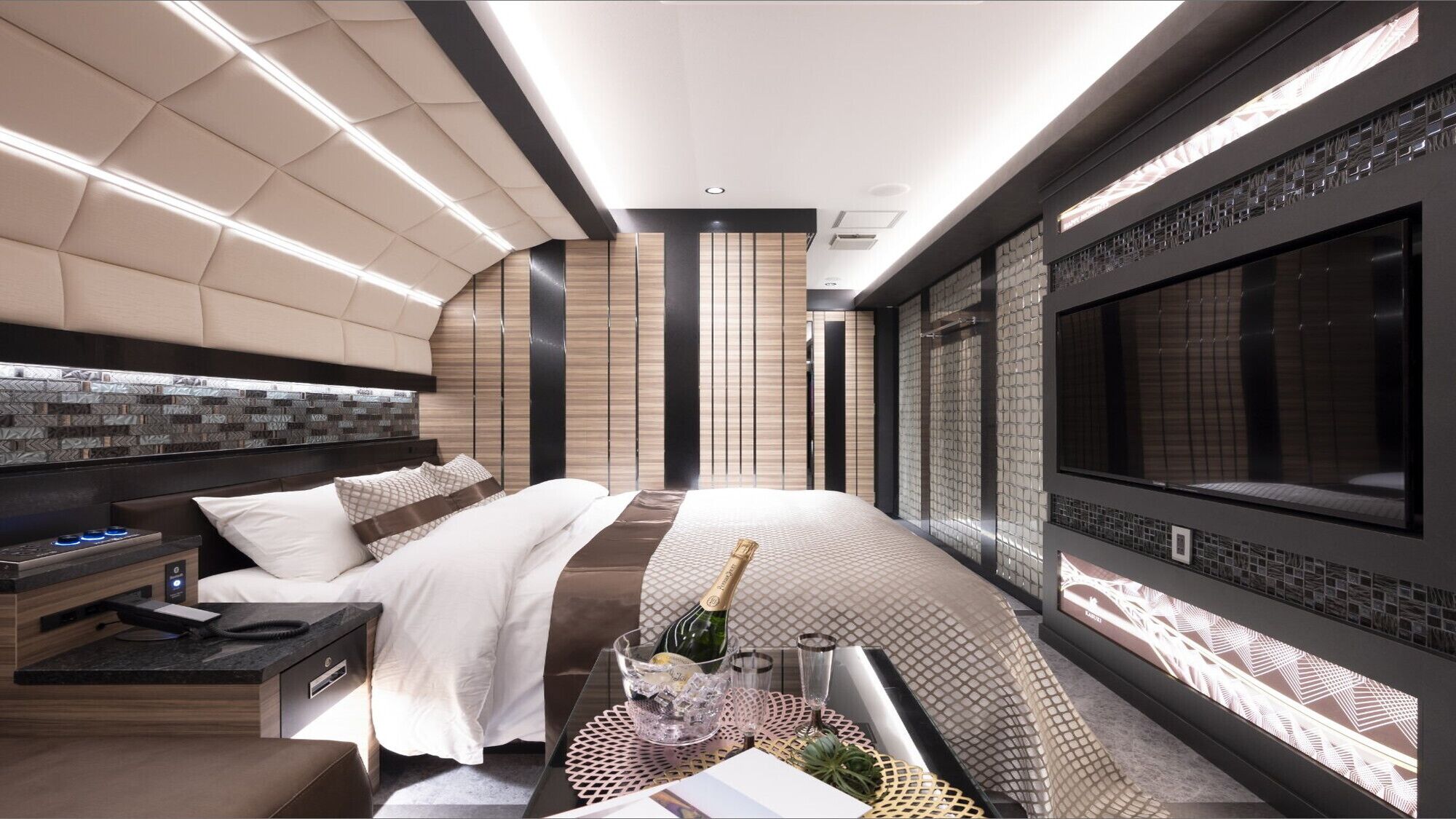 A modern hotel room with a bed, bedside table, minibar, and a large TV screen.