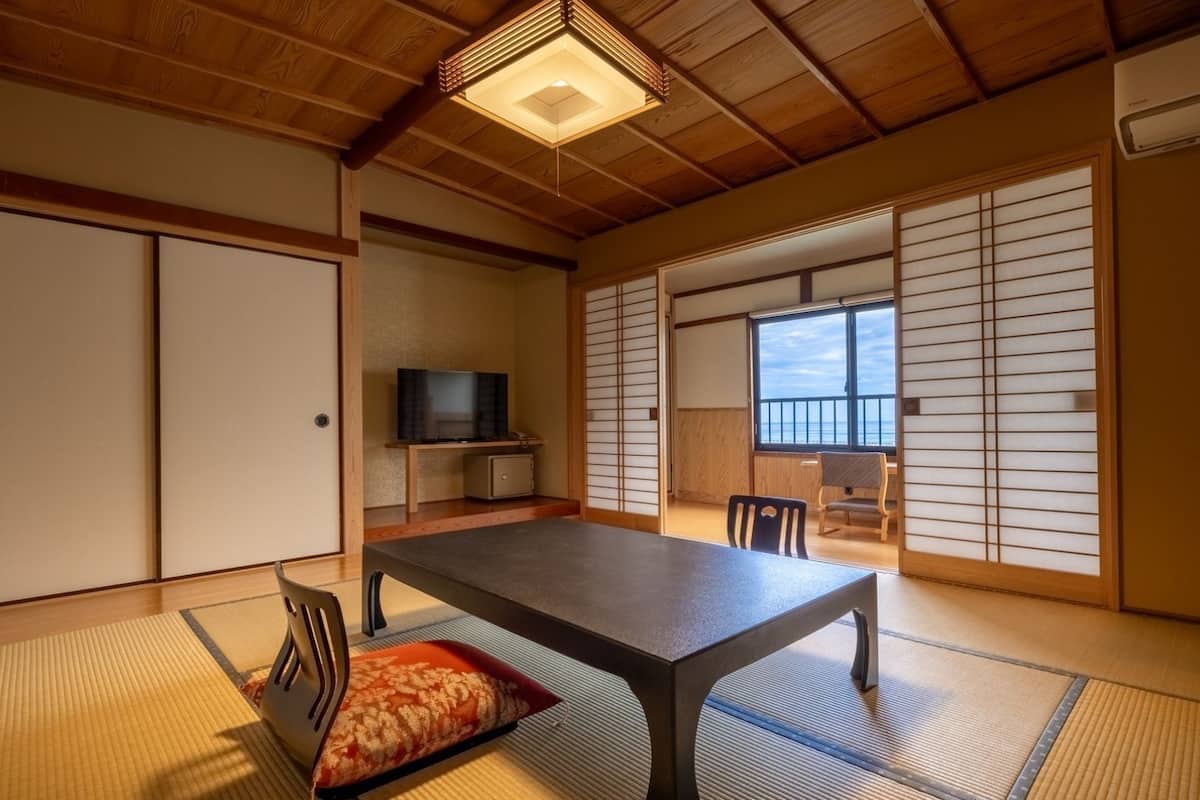 Ein traditioneller japanischer Raum mit Tatami-Böden, einem niedrigen Tisch, einem Fernseher und einer Schiebetür, die auf einen Balkon führt.