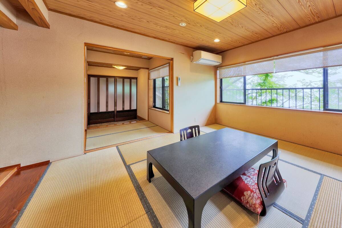 Ein Zimmer mit Tatami-Boden, einem niedrigen Tisch, einem Stuhl, einer Schiebetür und einem großen Fenster mit Aussicht.