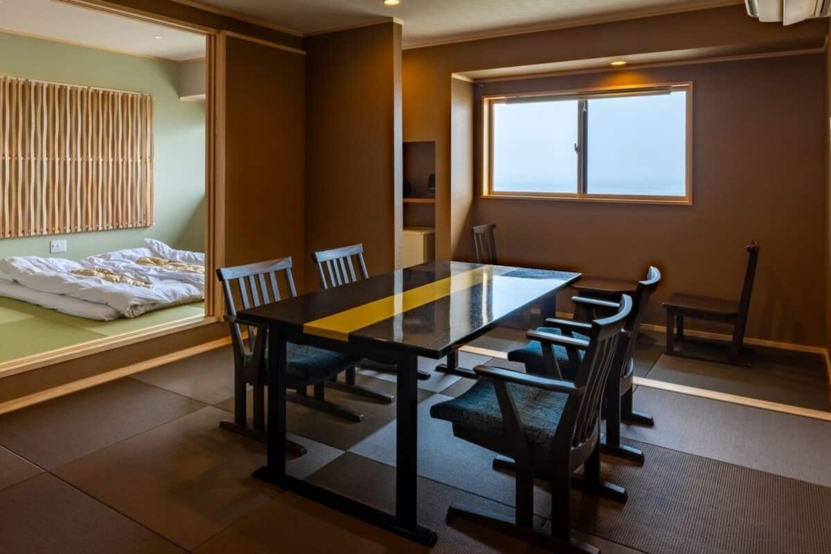 Ein Hotelzimmer mit Bett, einem Esstisch mit Stühlen, einem Fenster und Blick aufs Meer.