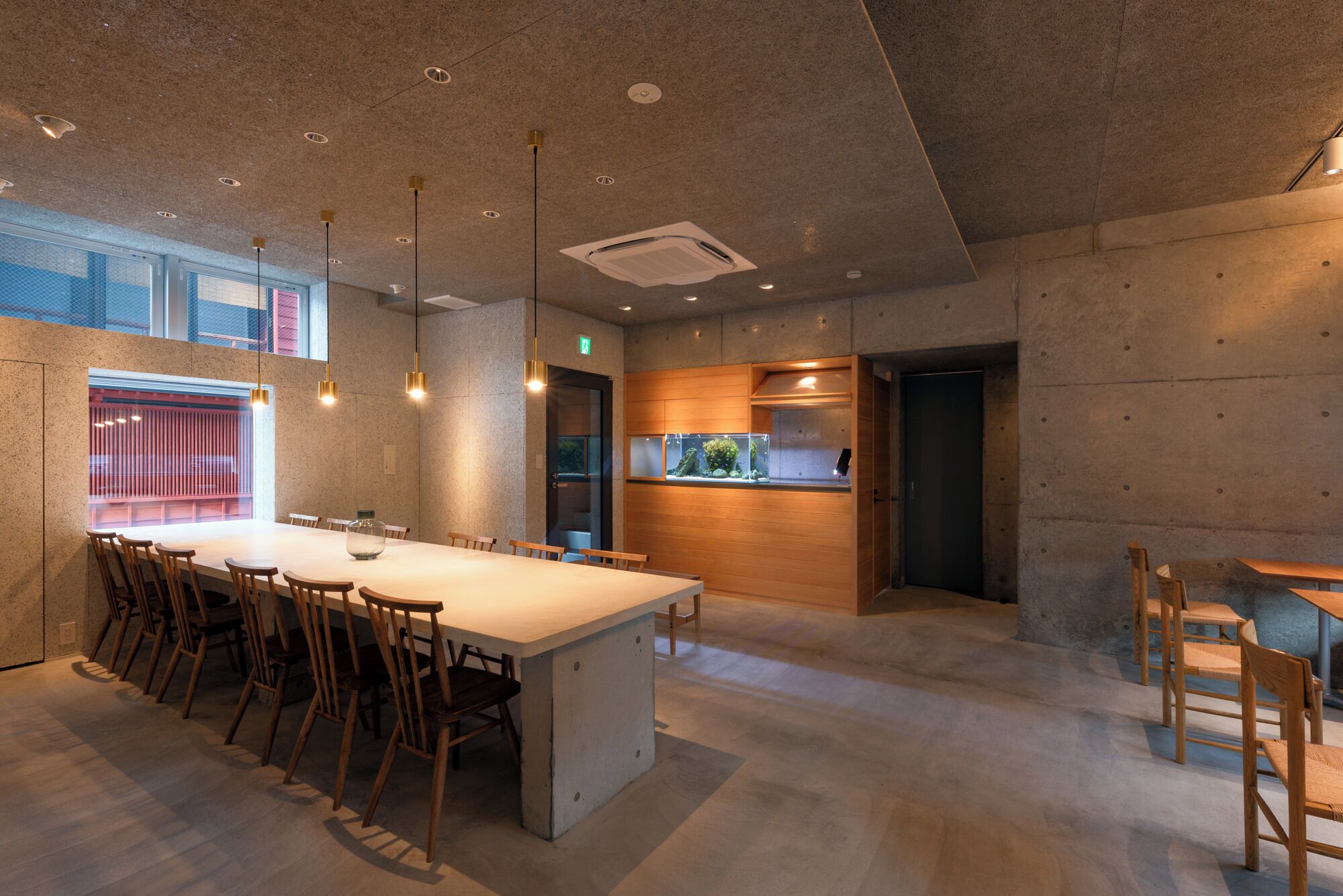 A modern dining area with a long table, chairs, pendant lighting, a fish tank, and a kitchen area with wooden cabinets.
