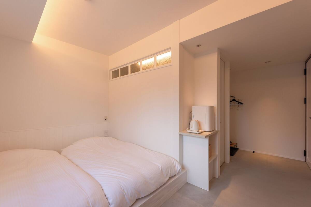 A bedroom with a bed, a bedside table with a lamp, a small shelf, and a closet with hanging clothes.