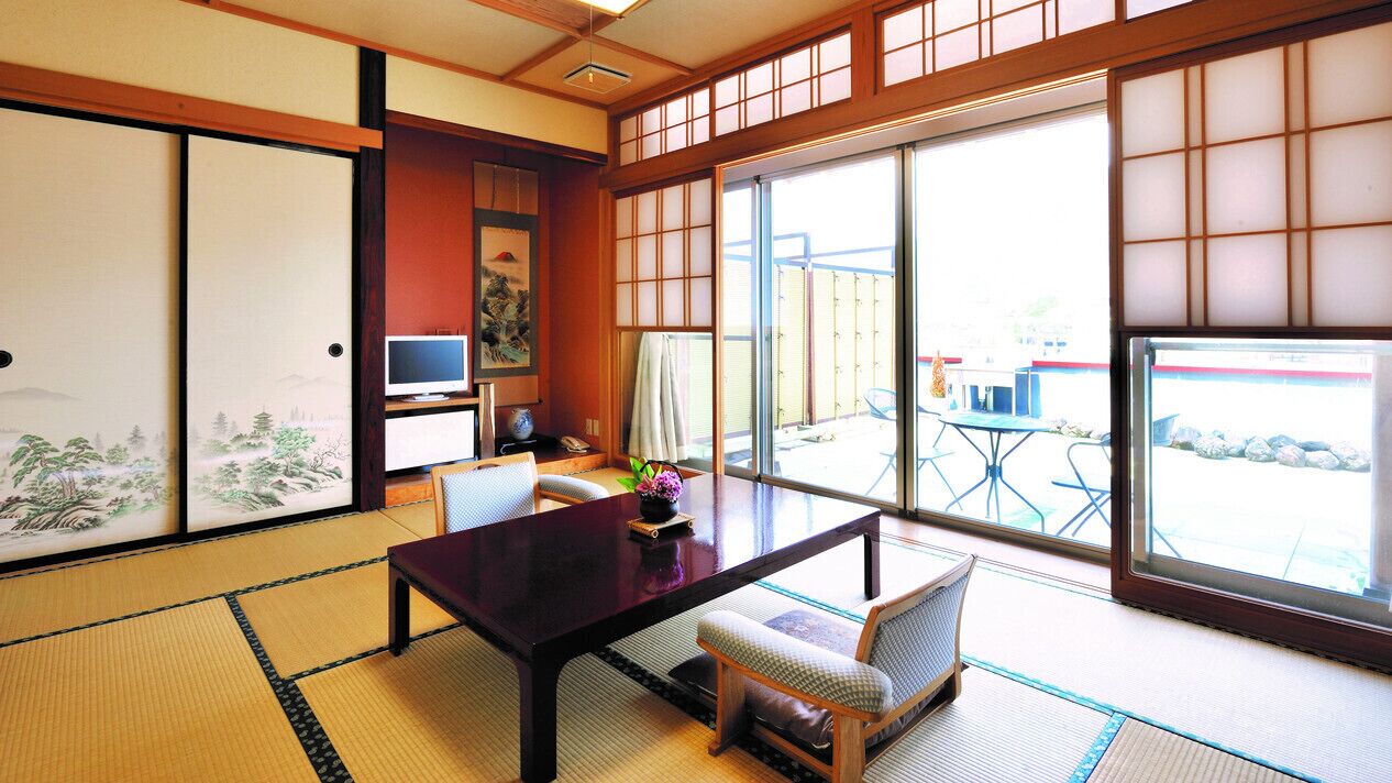 Una habitación japonesa tradicional con suelo de tatami, una mesa baja, un banco acolchado, puertas correderas con motivos decorativos y vistas al exterior.