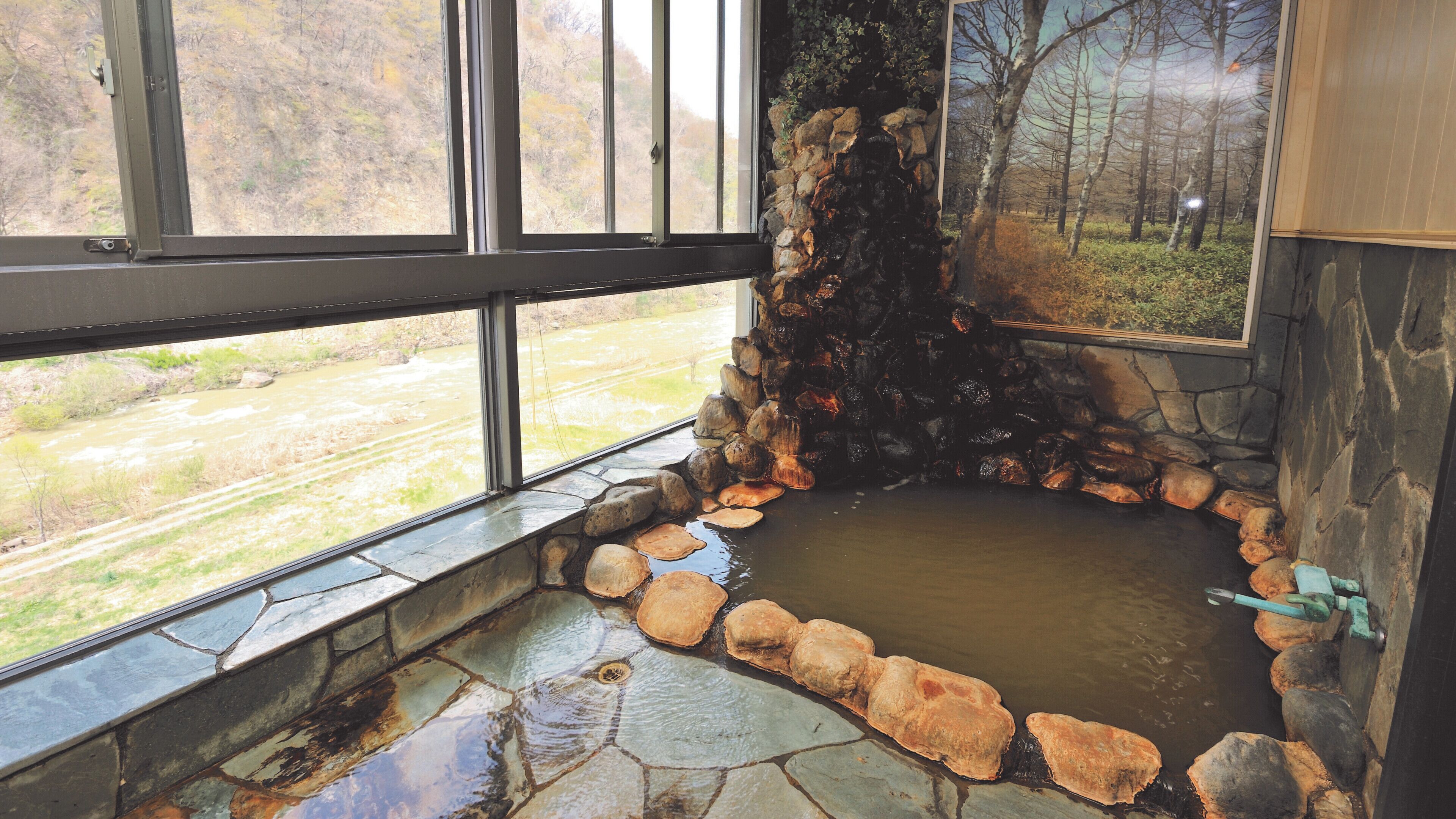 A stone hot tub with a view of a river and forest through large windows.