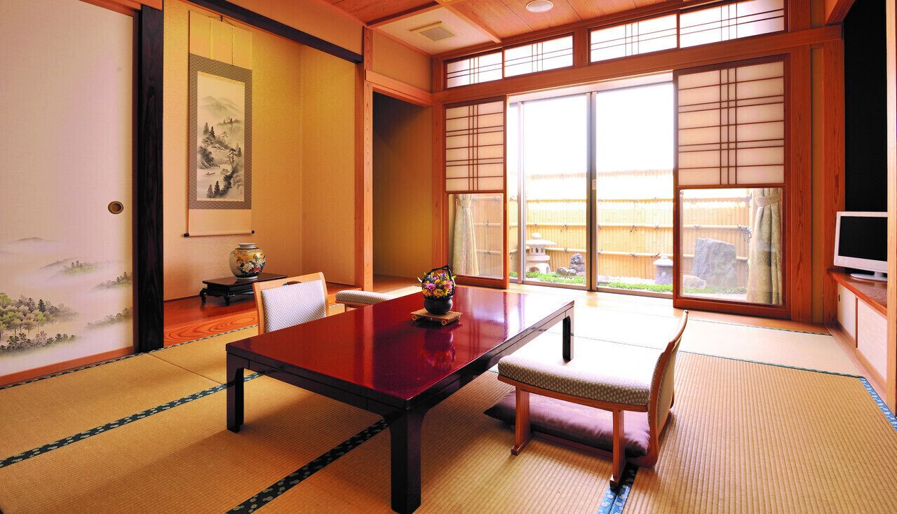 Una habitación japonesa tradicional con suelo de tatami, una mesa baja, una silla acolchada, un televisor y una puerta corredera con vistas al exterior.