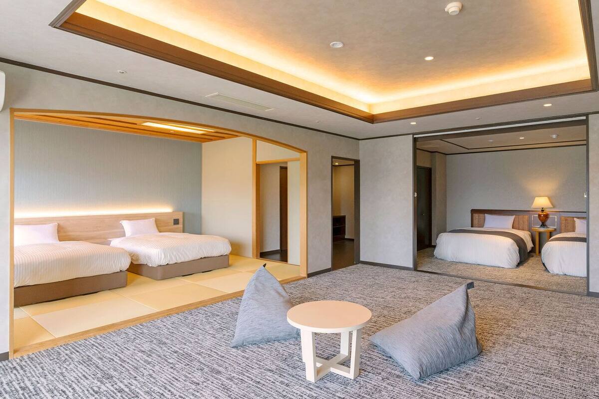 A modern hotel room with a large bed, a smaller bed, a seating area with a table, and a mirror reflecting the room.
