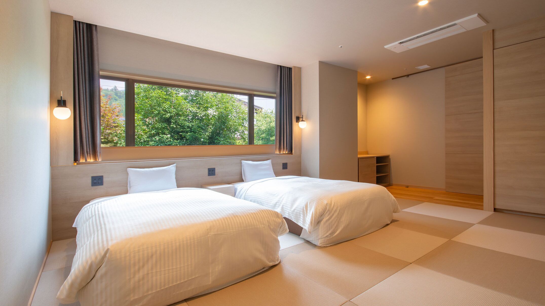 Quarto de hotel moderno com duas camas, uma janela grande e vista para área verde.