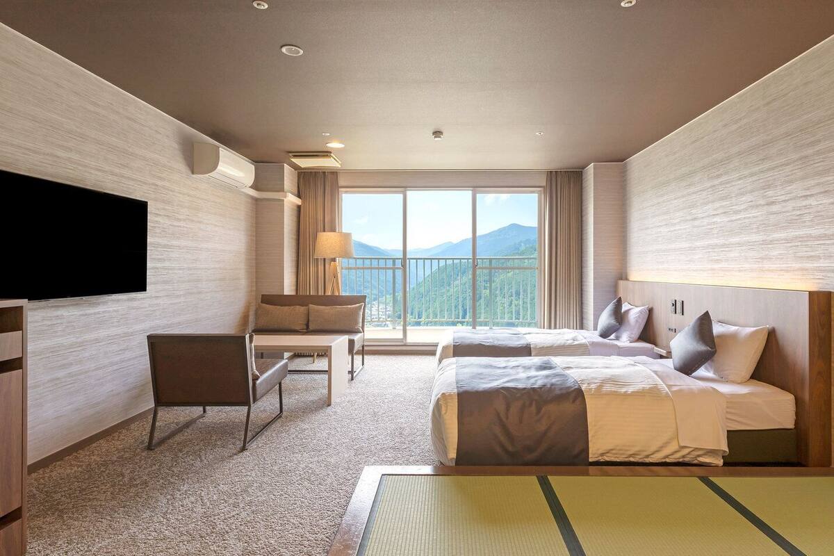 A hotel room with a TV, a desk, two beds, a chair, and a large window with a mountain view.