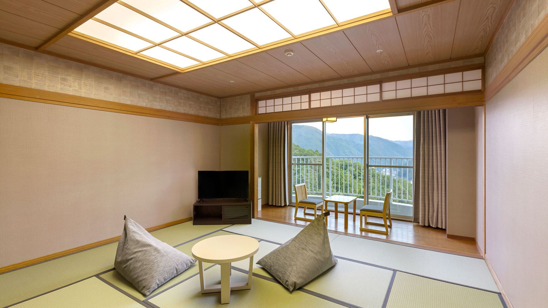 Quarto tradicional japonês com piso de tatame, televisão, mesa com cadeiras e varanda com vista para a montanha.