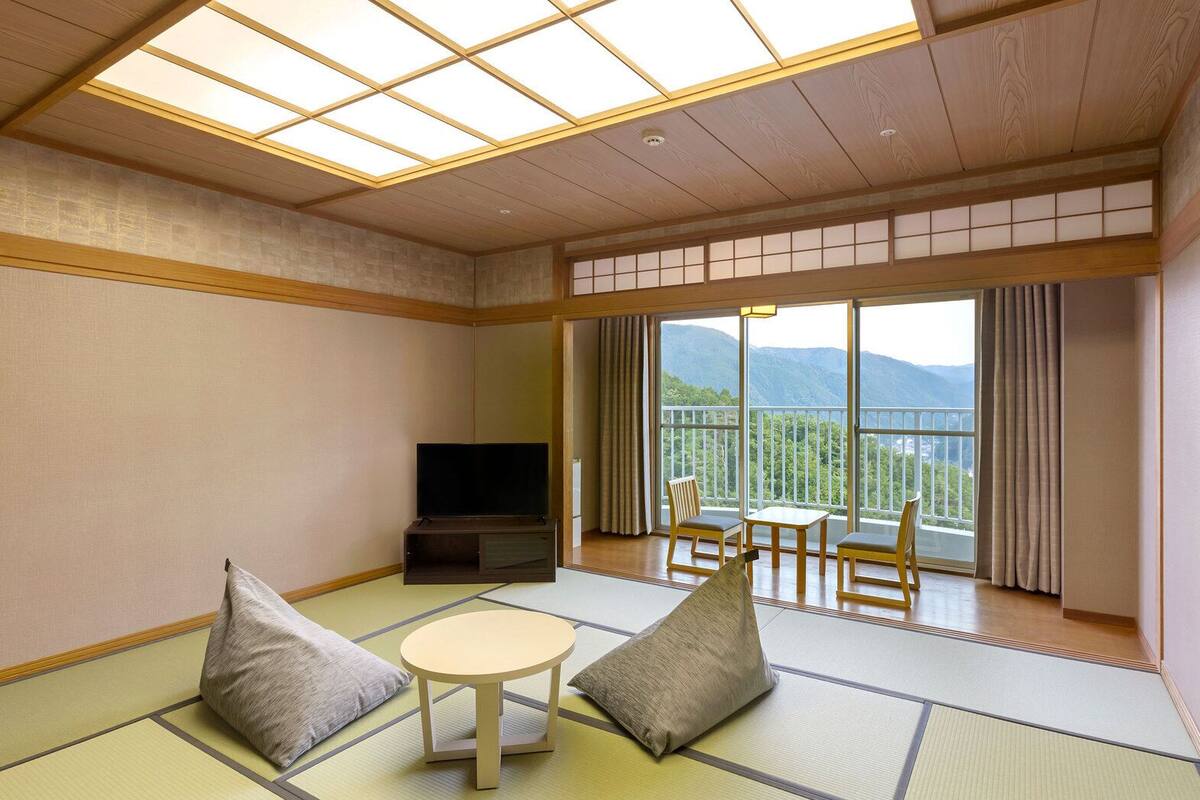 A traditional Japanese-style room with tatami flooring, a television, a table with chairs, and a balcony with a mountain view.