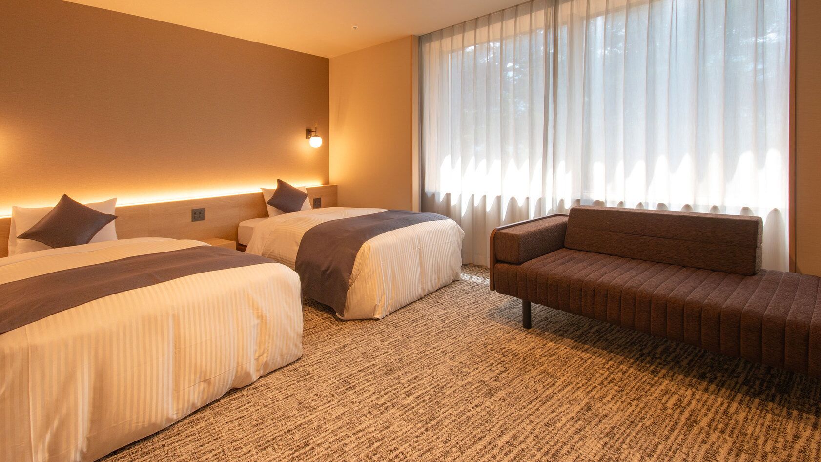 Quarto de hotel com duas camas, um sofá, uma janela grande com cortinas leves e um candeeiro fixado na parede.