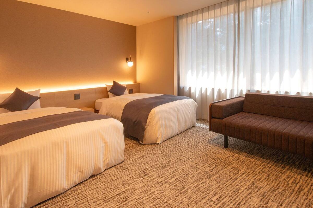 A hotel room with two beds, a sofa, a large window with sheer curtains, and a wall-mounted light fixture.