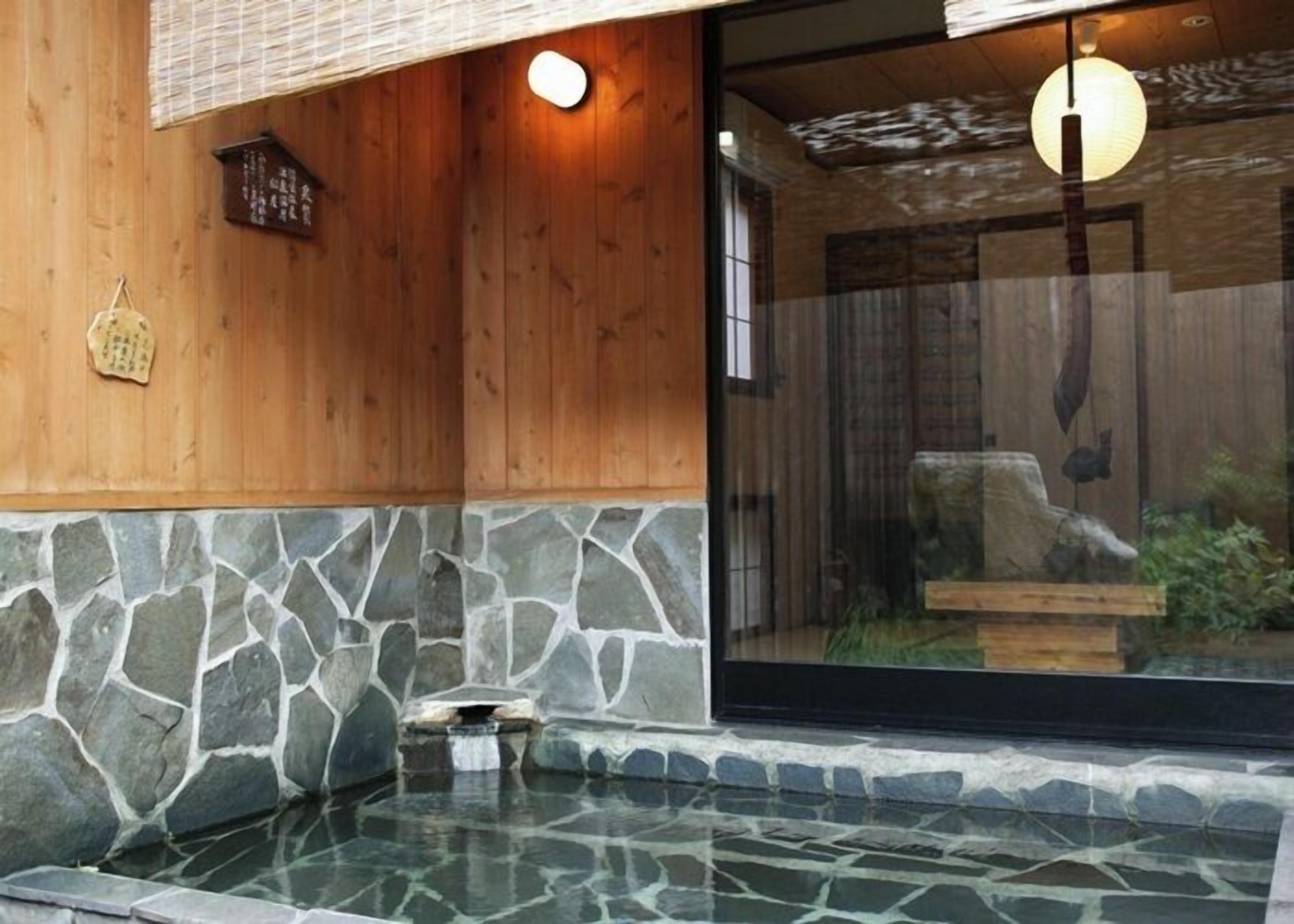 Ein Bereich mit Steinplatten-Whirlpool, Holzwänden und einer Glastür zu einem Raum im traditionellen japanischen Stil.