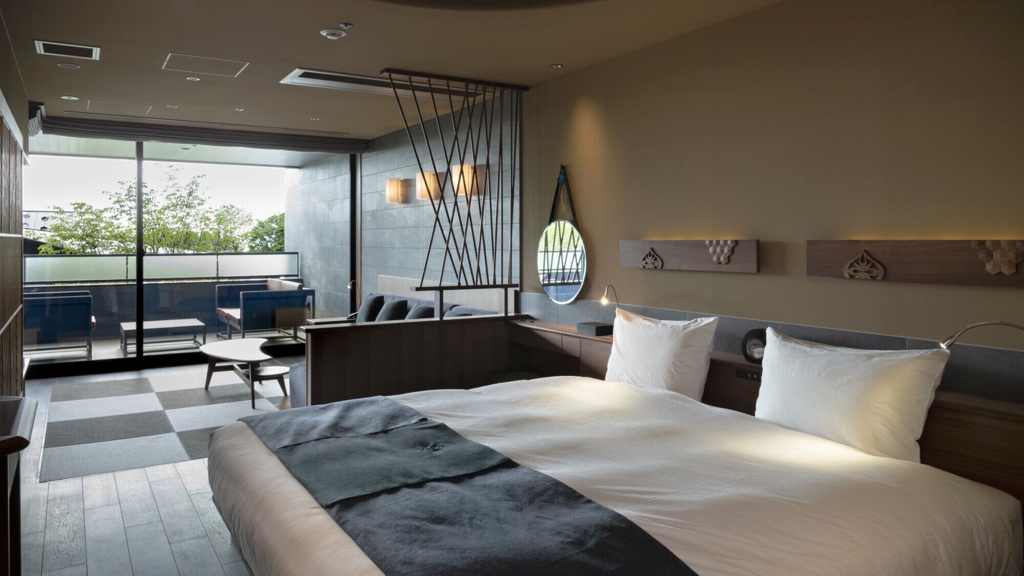 A modern hotel room with a large bed, a balcony with seating, and a view of the outdoors.