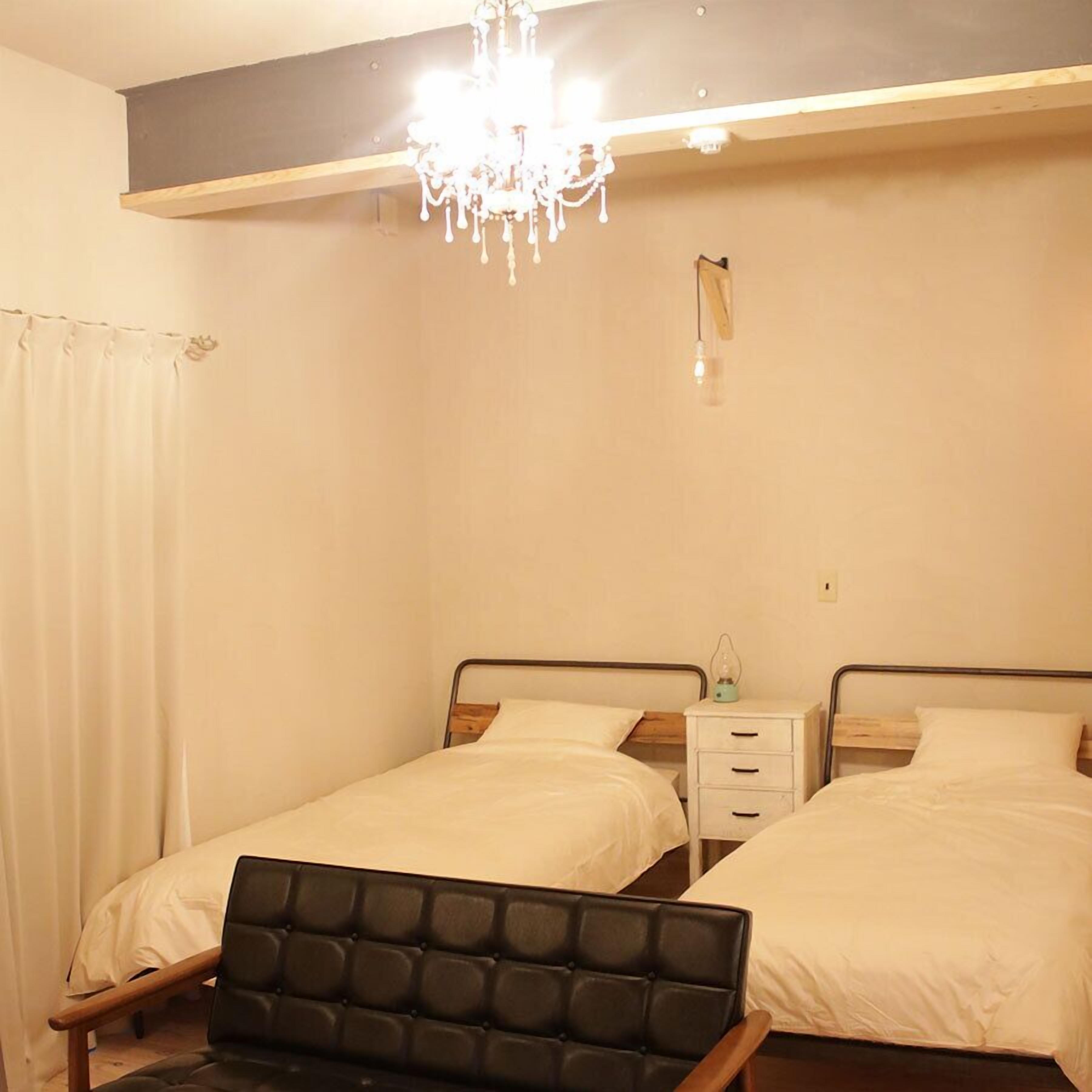 A bedroom with two beds, a nightstand with a lamp, a chair, and a chandelier.