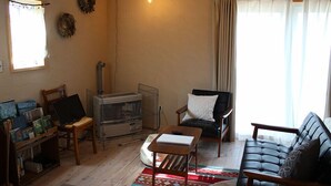 A living room with a sofa, armchair, and a small table. There is a window with curtains, a radiator, and a wall-mounted light fixture.
