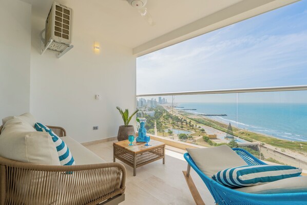 Ein Balkon mit Blick auf das Meer und die Stadtlandschaft, mit Korbmöbeln, einem kleinen Tisch und dekorativen Gegenständen sowie einer Klimaanlage.