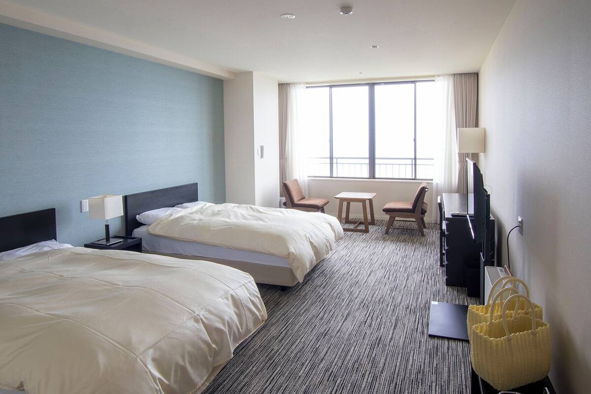 A hotel room with two beds, a desk, a chair, a TV, and a window with curtains.