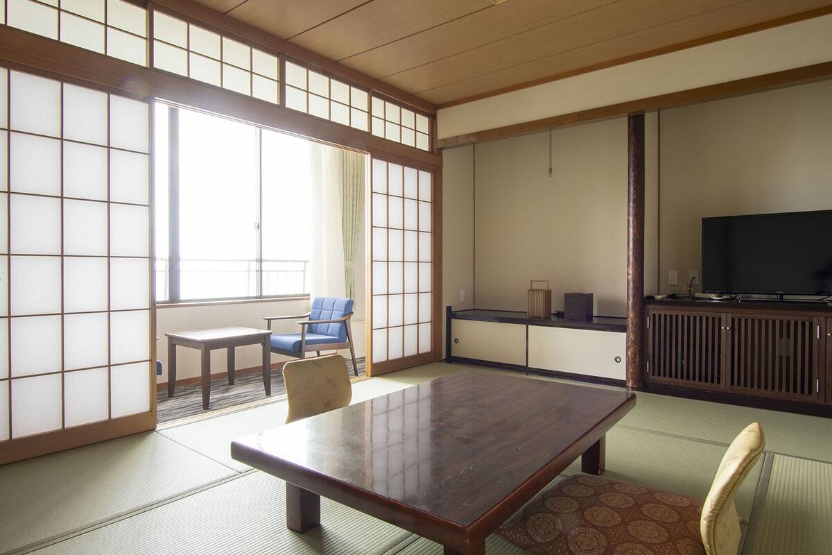 A traditional Japanese room with tatami flooring, a low table, a blue chair, and a television on a wooden stand.