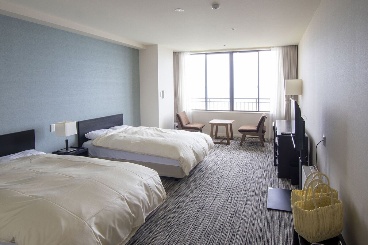 A hotel room with two beds, a desk, a chair, a TV, and a window.