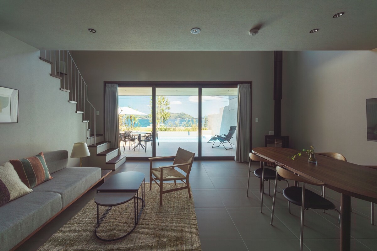 Un soggiorno moderno con divano, tavolino da caffè e tavolo da pranzo. Una scala conduce a un balcone con vista sul mare.